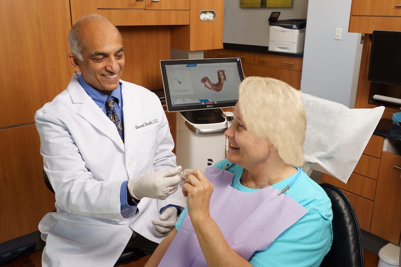 Dr. Sharad Pandhi, wearing a white coat and gloves, smiles while handing a dental sealant to a female patient wearing a bib in a modern dental office. A digital dental scan is visible on the screen behind them.