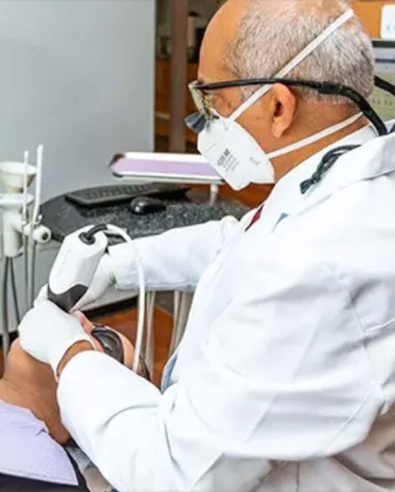 A dentist wearing a white coat, protective glasses, and a face mask takes a panoramic digital x-ray of a patient's teeth using an intraoral scanner in a modern dental clinic.